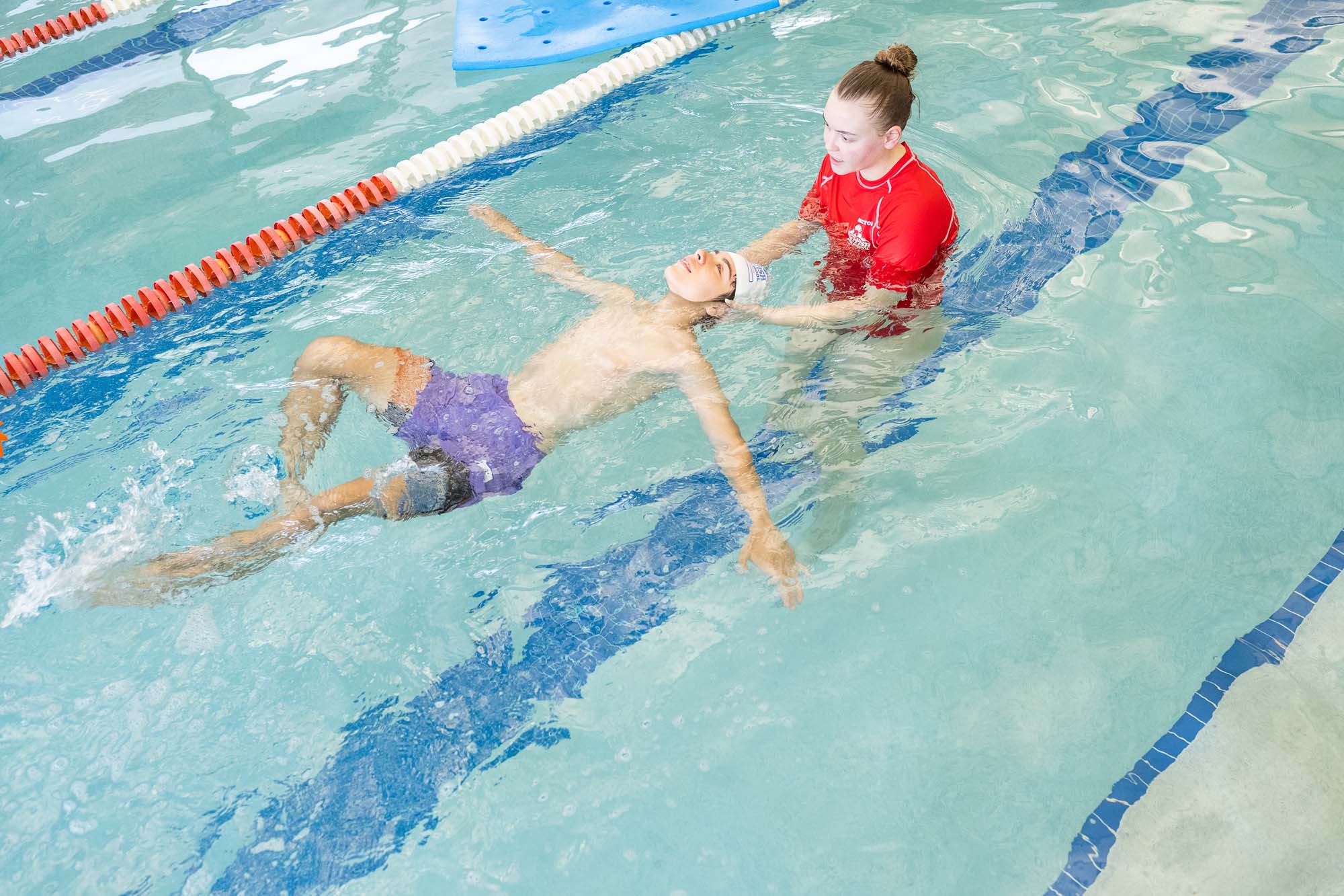 A swim instructor helps a young boy practice floating on his back in a swimming pool.