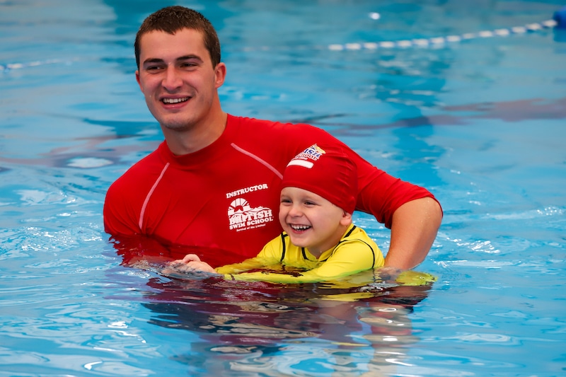 A smiling swim instructor helps a happy child learn to swim in a bright blue pool.