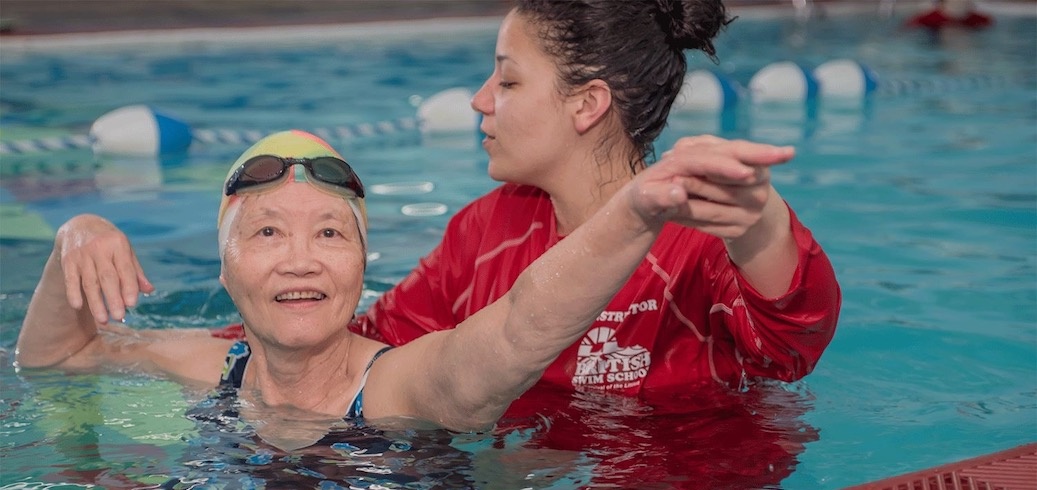An older woman in a swim cap smiles while a swim instructor helps her in the pool