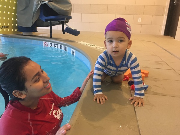 A swim instructor smiles at a baby in a swim cap crawling by the pool.