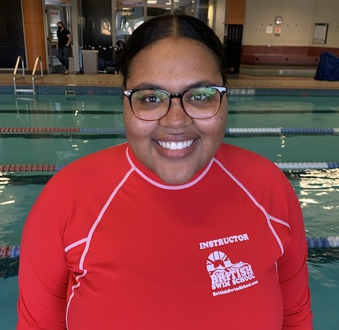 Smiling swim instructor in a red rash guard stands by an indoor pool.