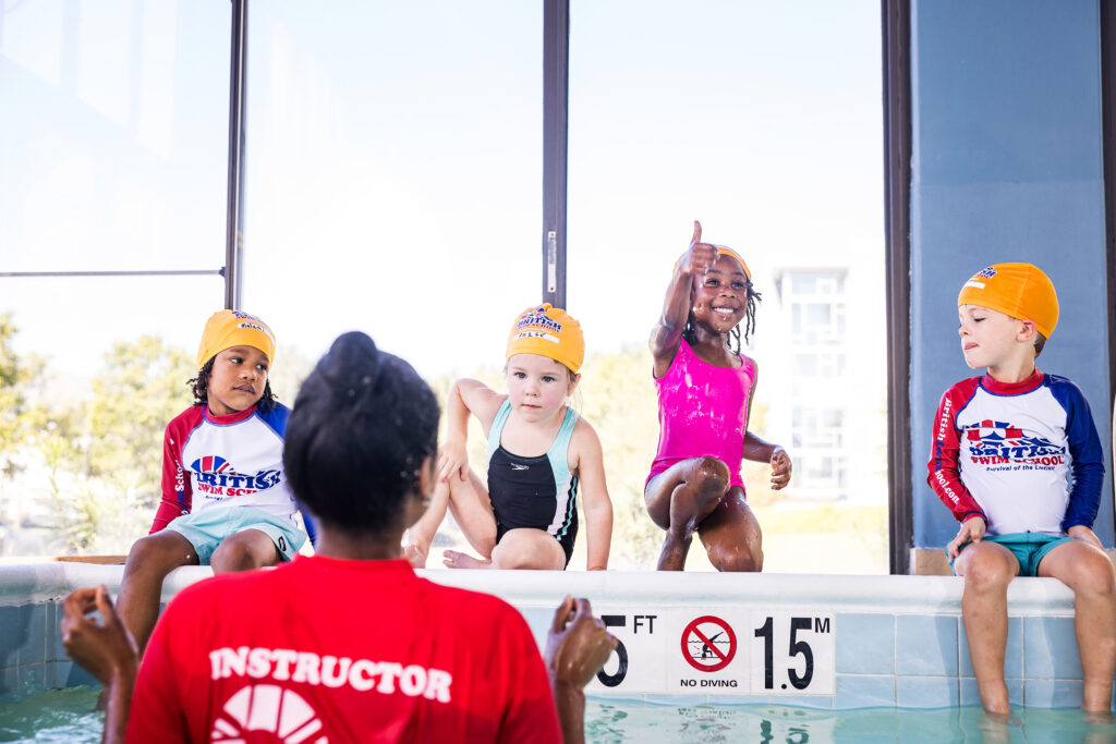 4 young children wearing orange British Swim School caps sit on the edge of an indoor pool during a swim lesson. A swim instructor faces the group while one girl gives an enthusiastic thumbs-up.