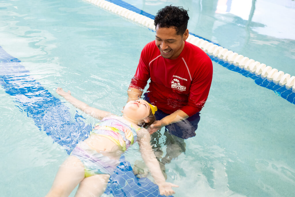 A British Swim School instructor supporting a toddler floating on their back during a swim lesson in Austin.