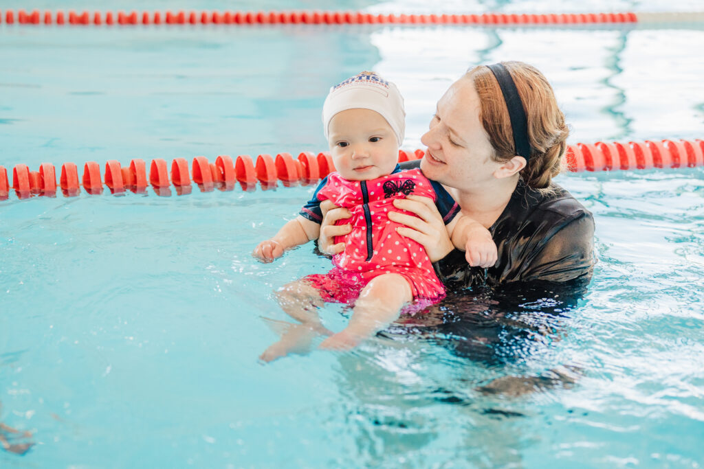 A smiling mother holds her baby in an indoor pool during infant swim lessons at British Swim School of Austin.  