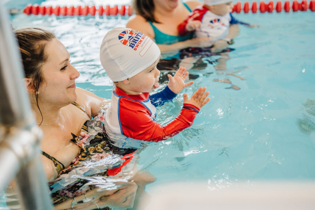 A parent holding their baby wearing a British Swim School swim cap during an infant swim lesson in Austin, with other caregivers and babies visible in the background.