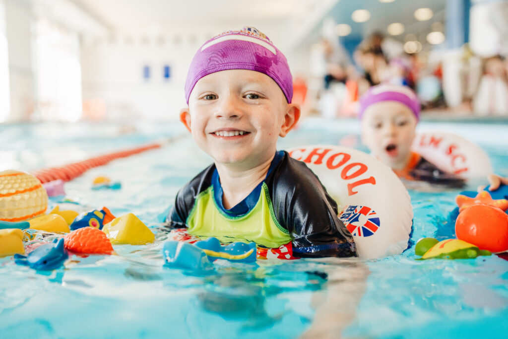 Young child in a British Swim school swim cap and pool floatie smiling during an indoor swim lesson.