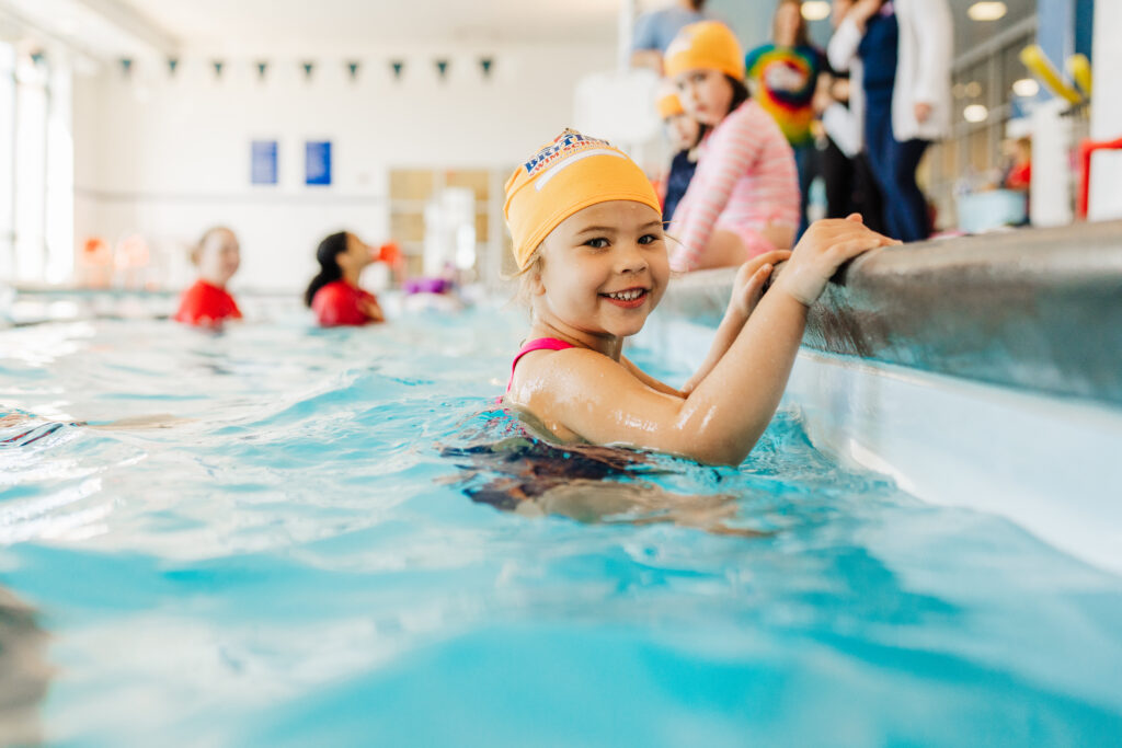 A young child at smiling as she holds on to the pools edge in an indoor heated pool at British Swim School