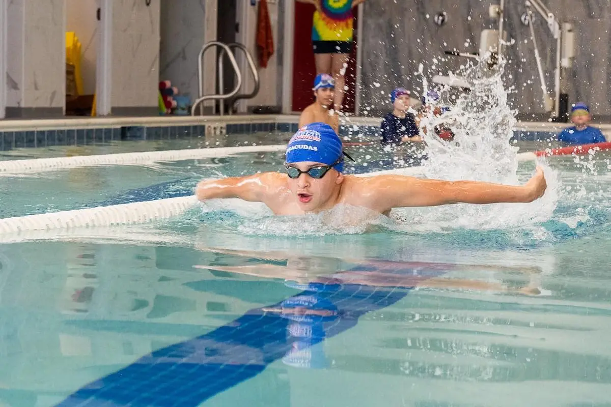 A young male swimmer in a blue cap performs the butterfly stroke, splashing water in a pool.