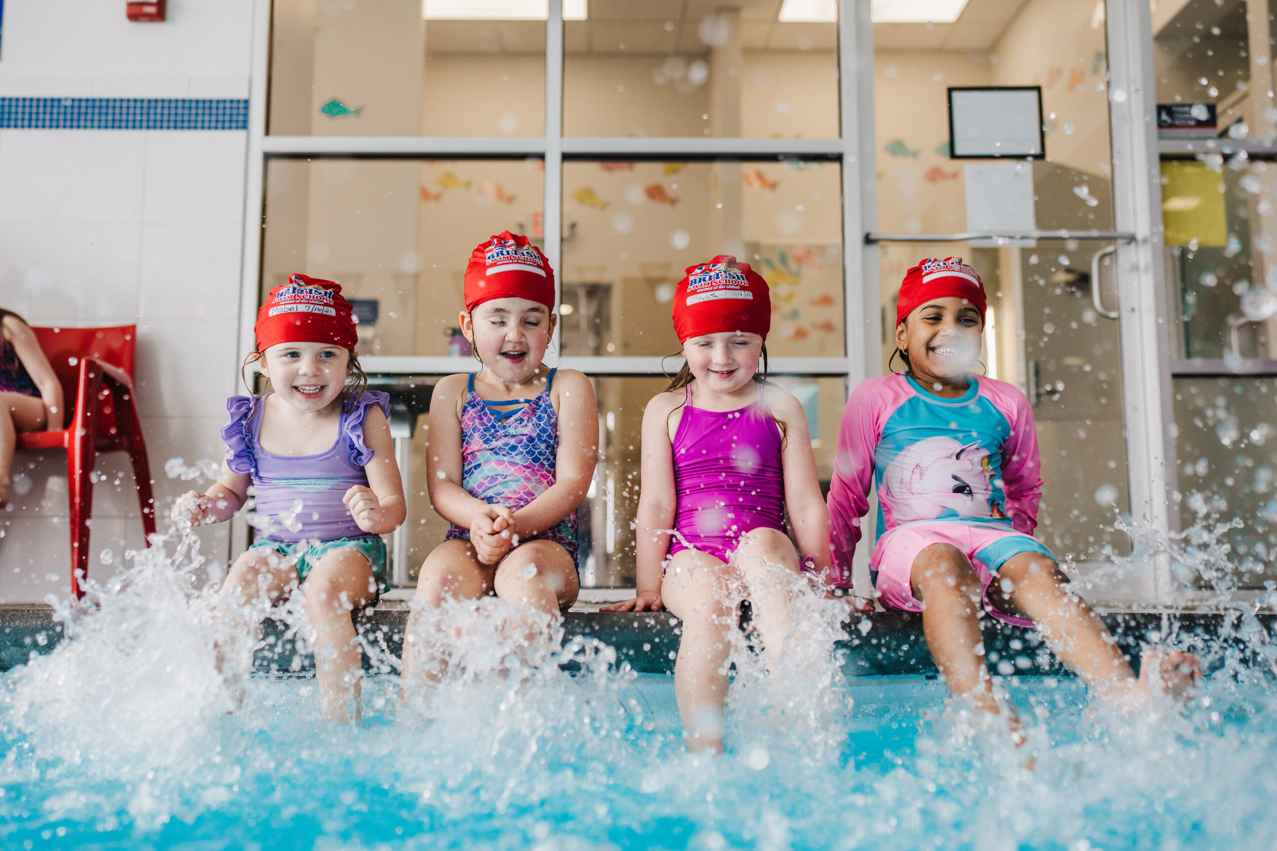 Four happy children wearing red British Swim School caps sit on the edge of an indoor pool, kicking their feet and creating a large water splash.