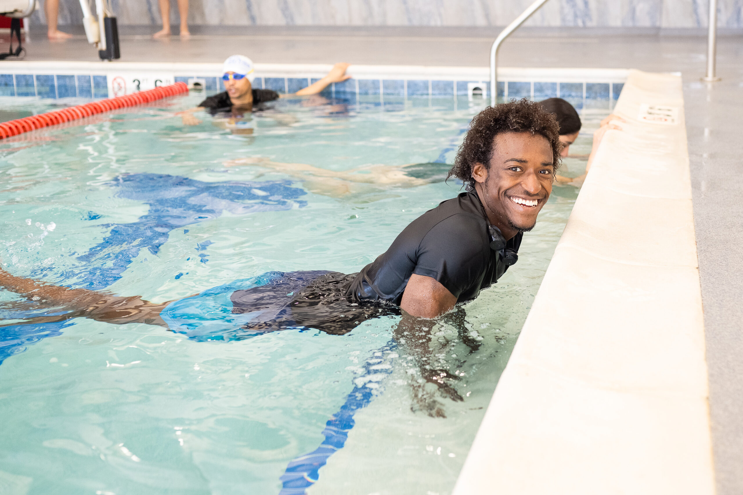 A smiling adult swimmer in a black swim shirt holds the edge of an indoor British Swim School pool during a lesson, with other swimmers practicing in the lane behind him.