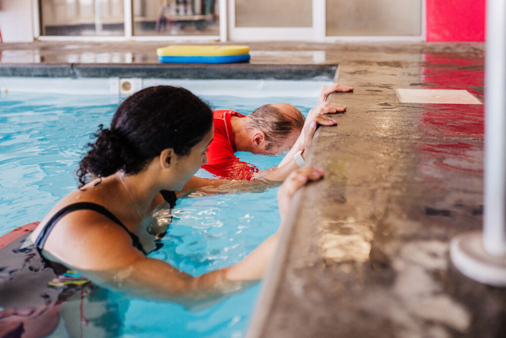 An adult student in a black top stays close beside a British Swim School instructor as they demonstrate holding the wall and lowering their face into the water at an indoor heated pool.
