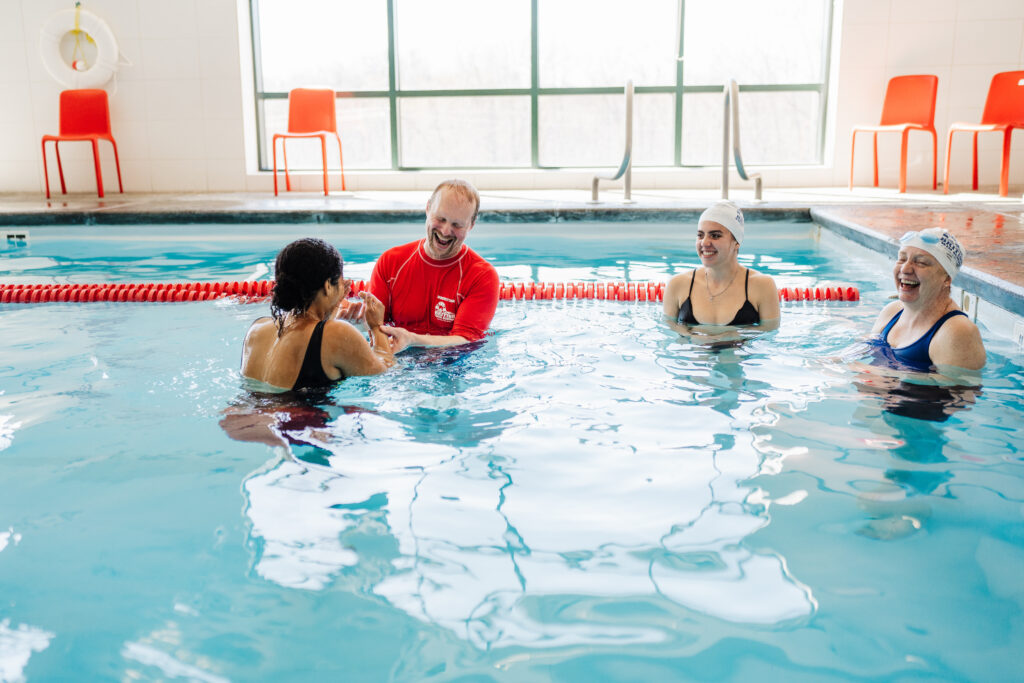 A British Swim School instructor in a red shirt leads a small group adult swim lesson, laughing with three adult students standing in an indoor pool.
