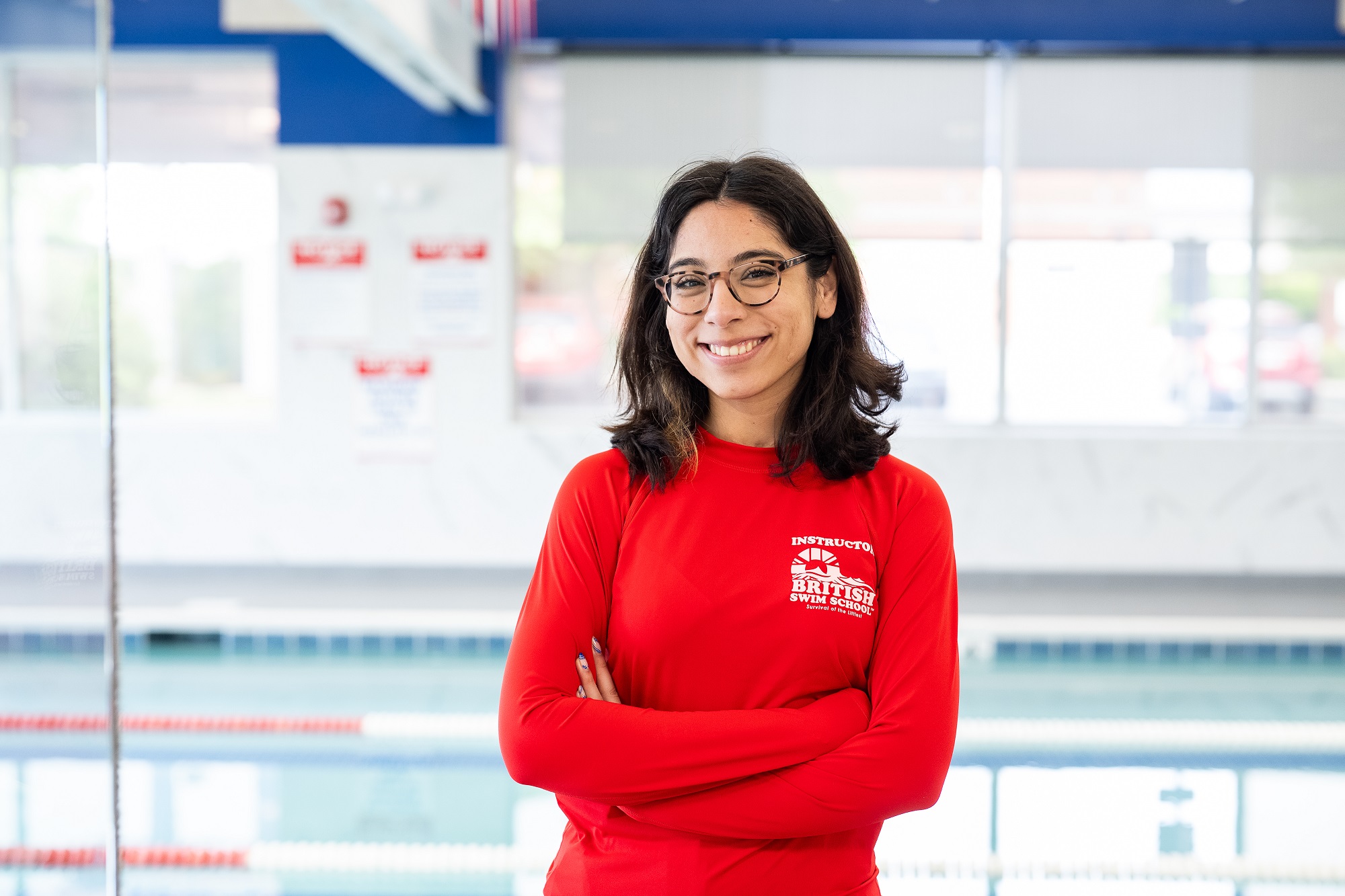 Smiling British Swim School instructor in a red shirt stands with arms crossed by a pool.