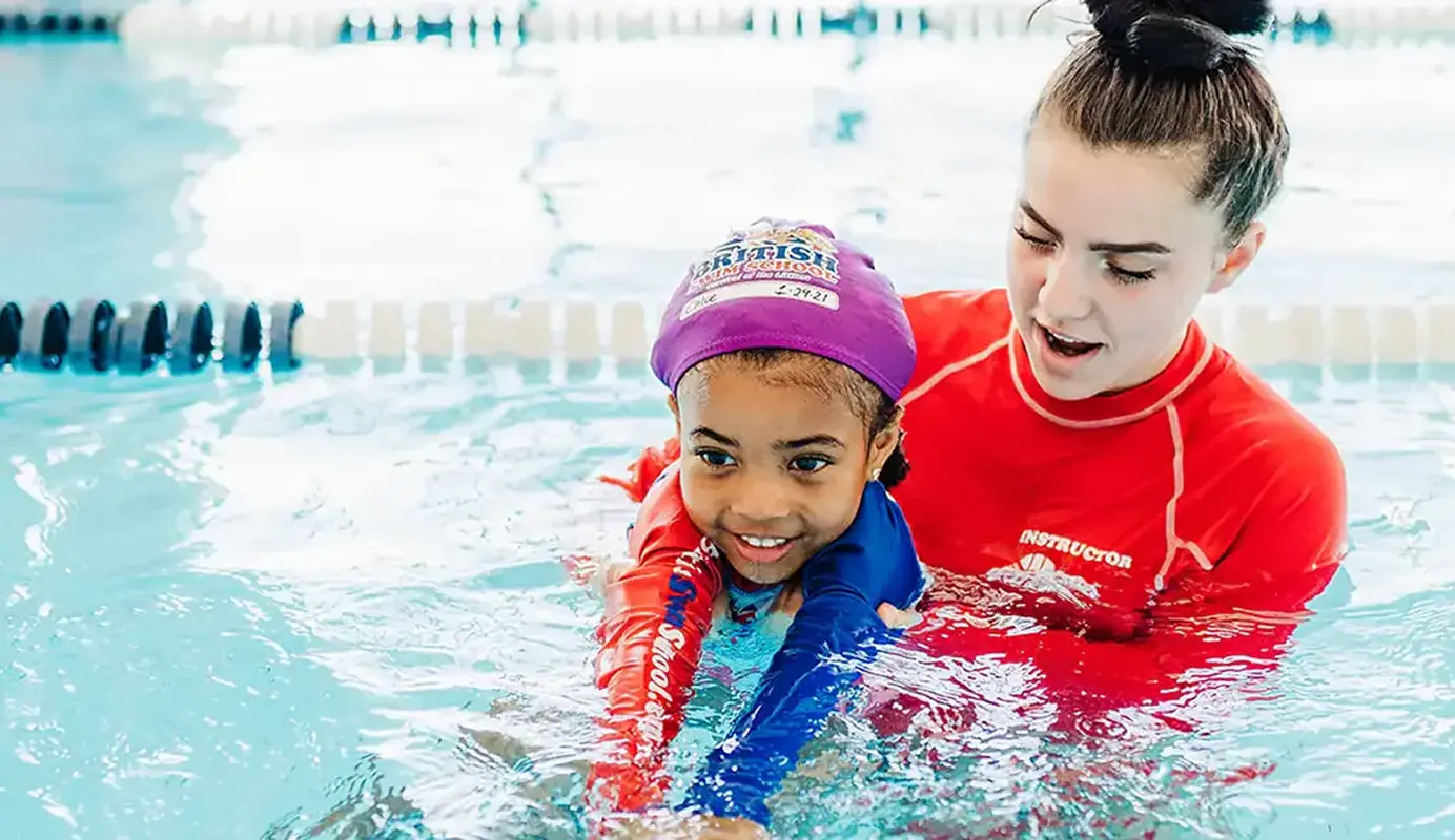 A smiling child in a purple swim cap learns to swim with an instructor's help.