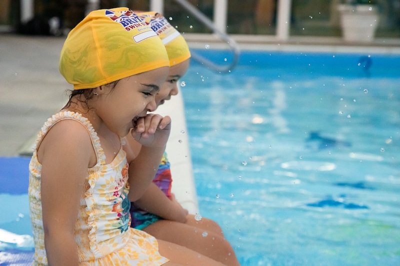 Two young girls in yellow swim caps sit by a blue swimming pool with water splashing