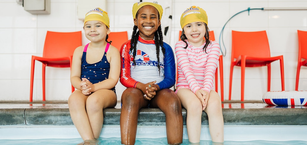 Three smiling girls in swimsuits and yellow caps sit by a pool.
