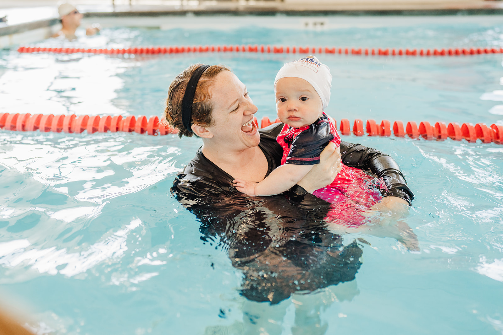 A smiling woman gently holds a baby wearing a swim cap in a pool.