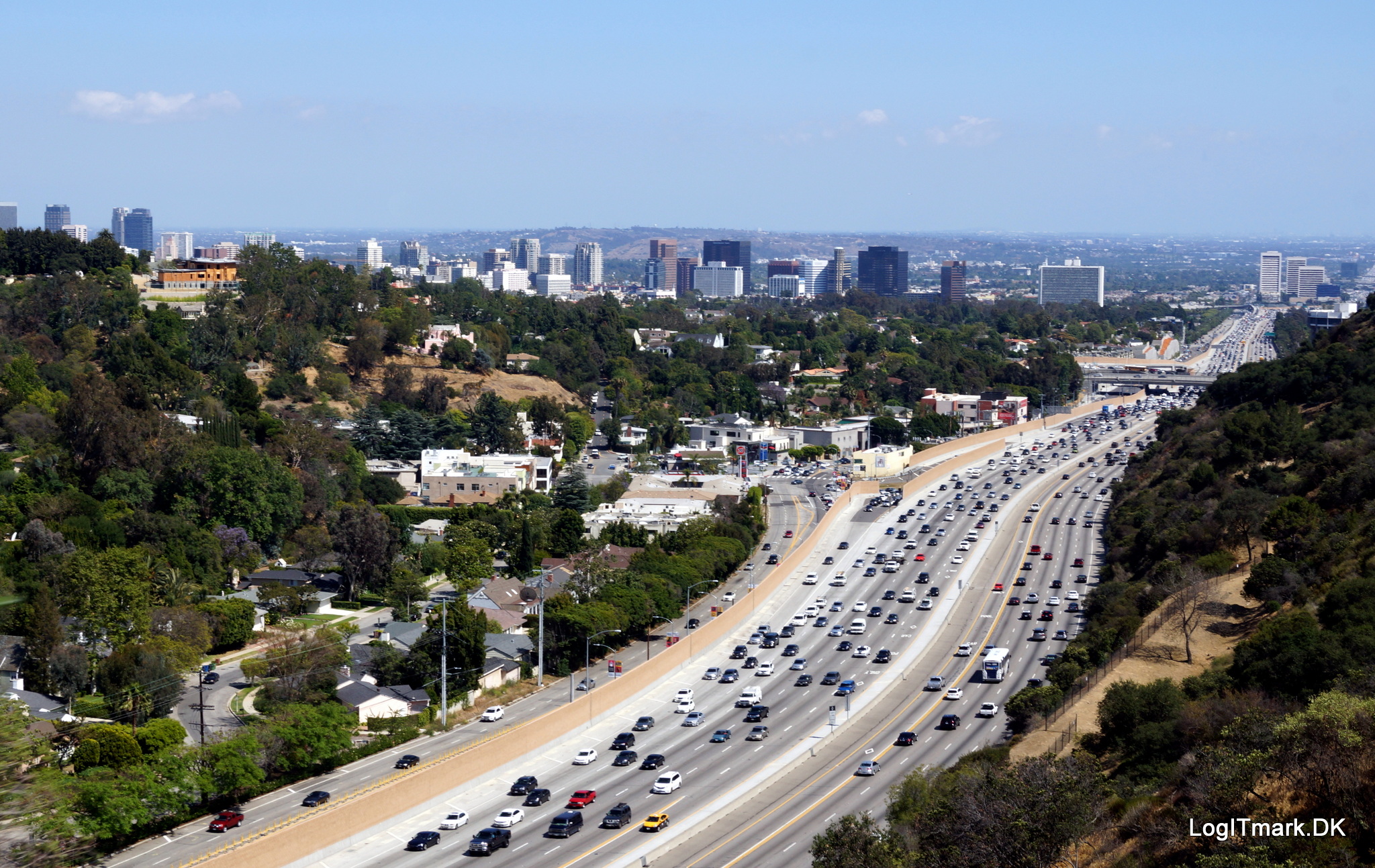 Heavy traffic flows on a wide freeway surrounded by lush hills and urban buildings under a