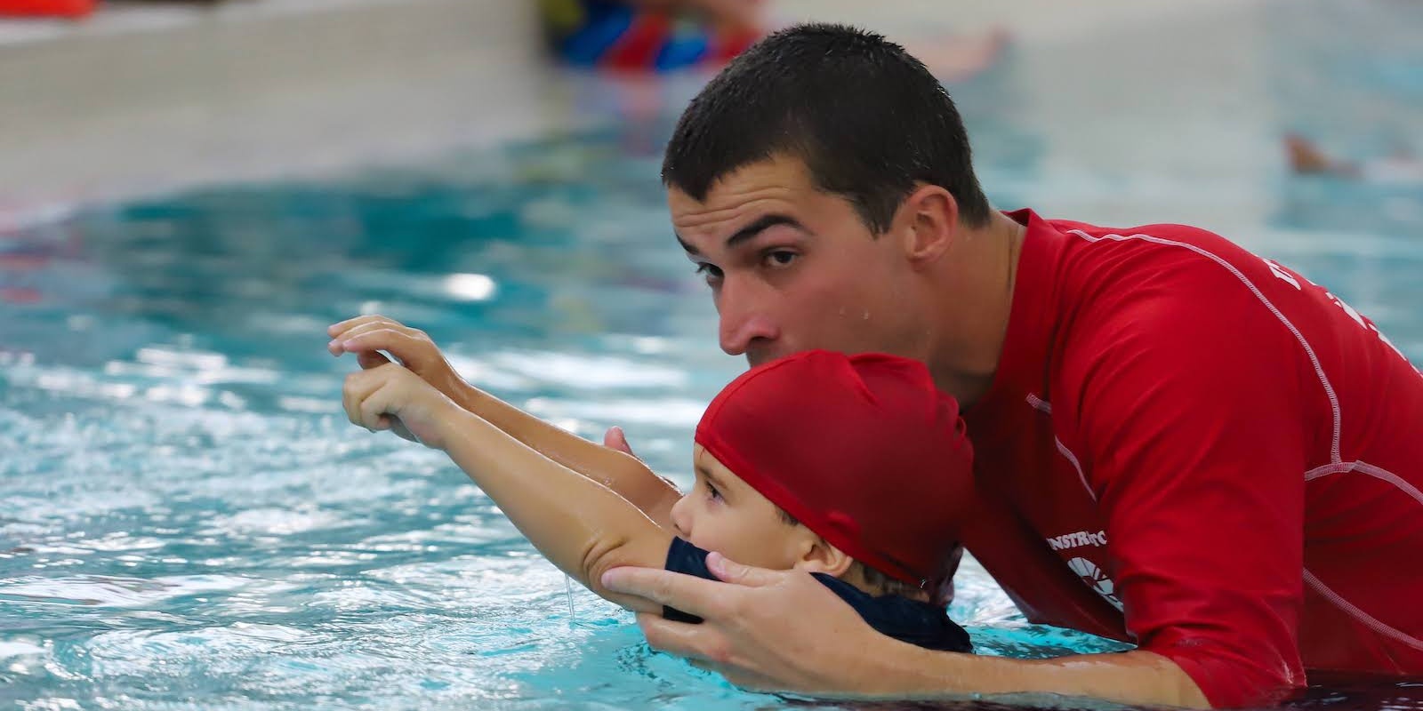 A swim instructor guides a child in a red cap to extend their arms while learning to