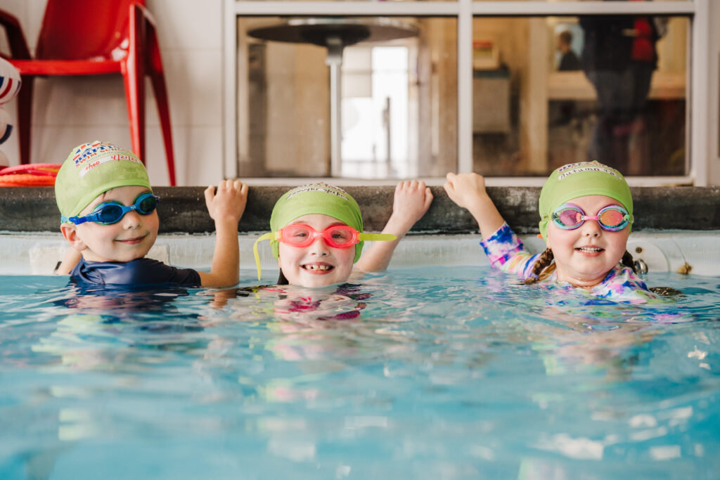 Three children wearing swimming gear smile from the edge of an indoor pool at British Swim School in Valley Spring, NY.