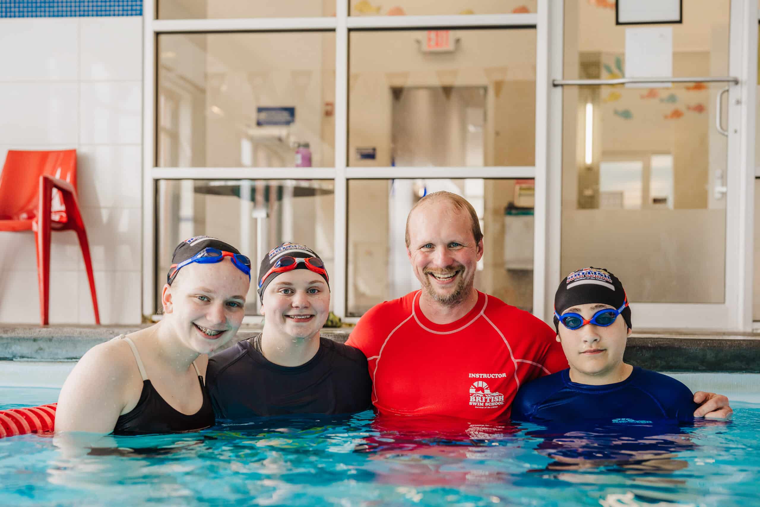A happy swim instructor and three students pose together in a bright indoor pool.