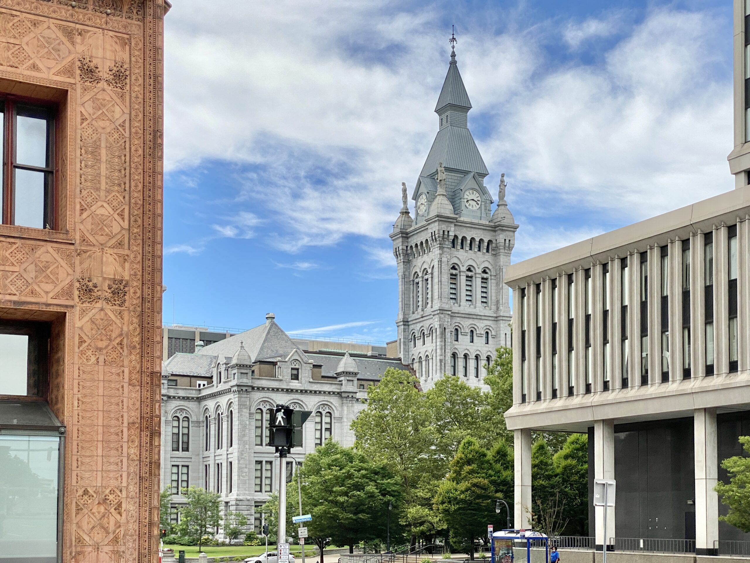 A historic grey clock tower dominates a city scene with diverse architectural styles under a cloudy blue