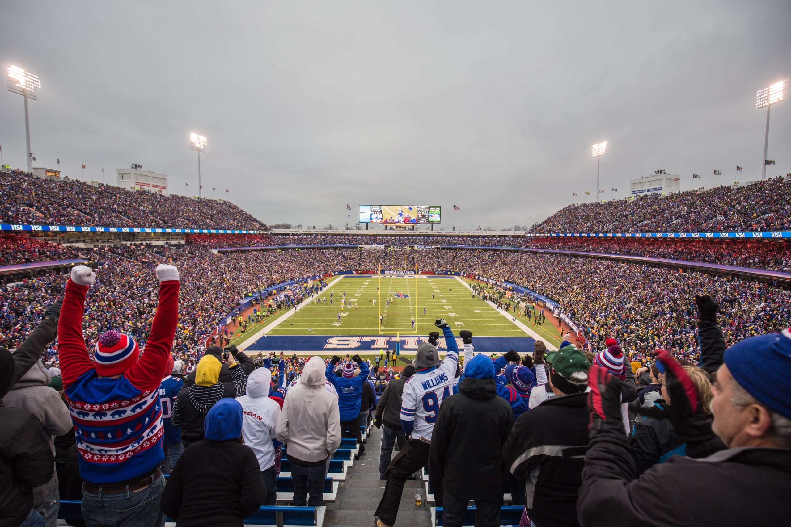 Cheering fans fill a large football stadium, watching a game on the field below.