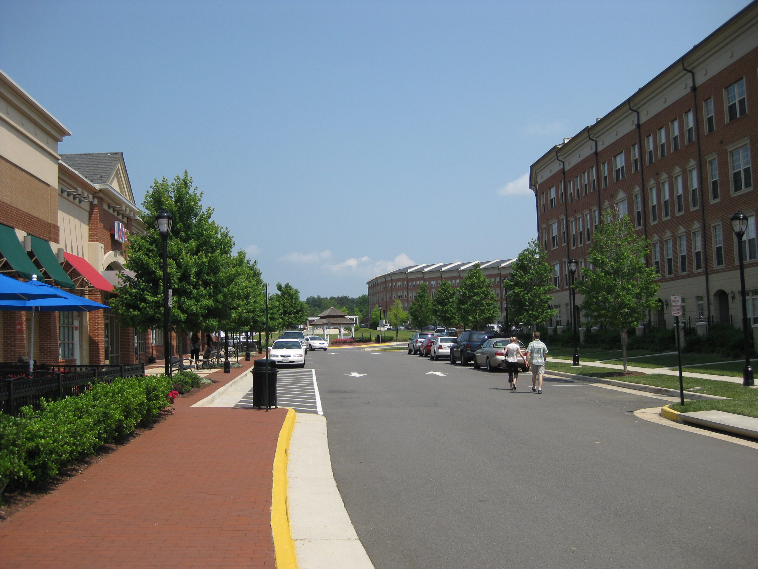 Pedestrians walk down a sunny street lined with brick buildings, shops, and
