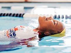 A smiling swimmer wearing a yellow cap floats on their back in a blue pool. TITLE