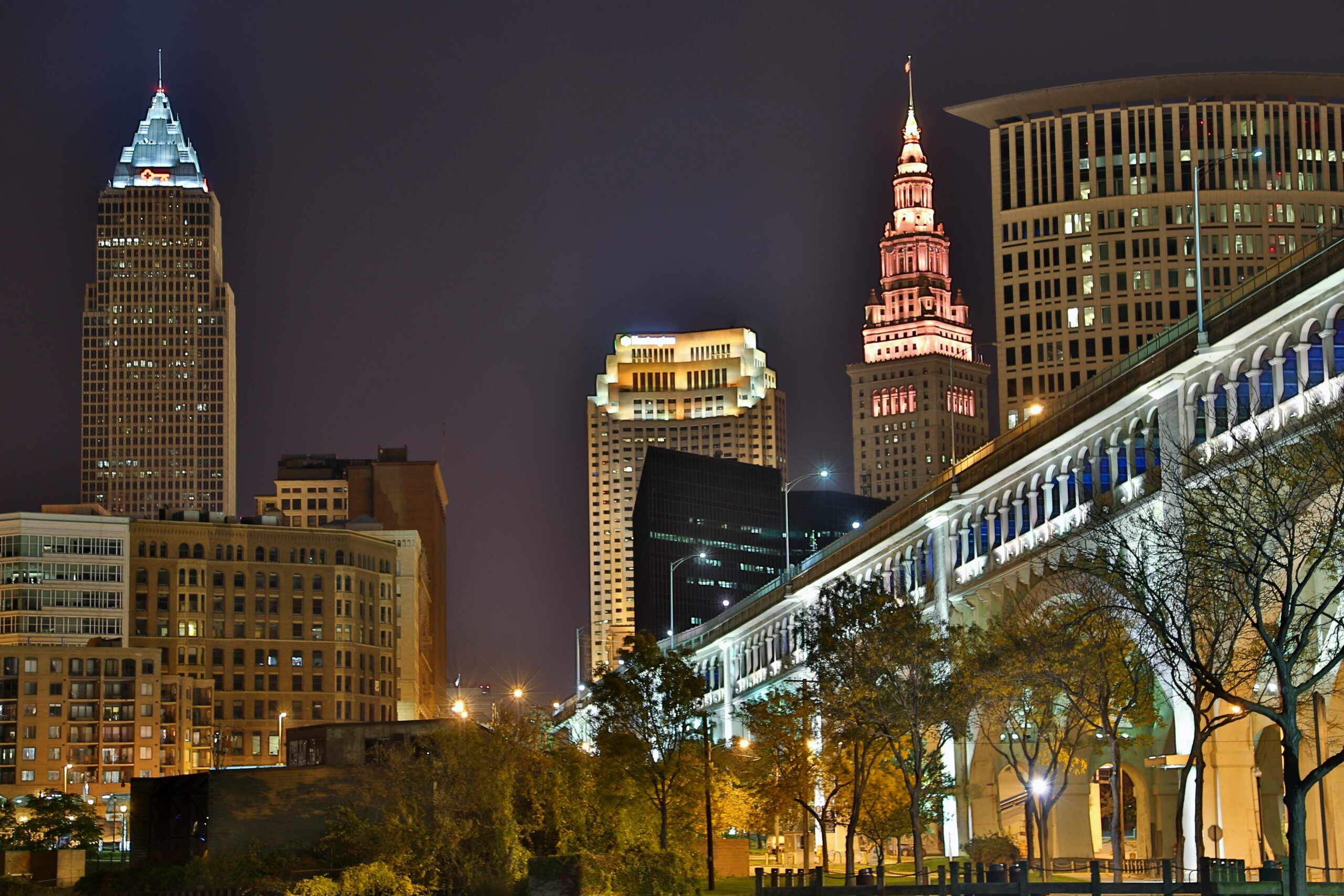 The Cleveland city skyline glows at night with iconic skyscrapers and a well-lit bridge.
