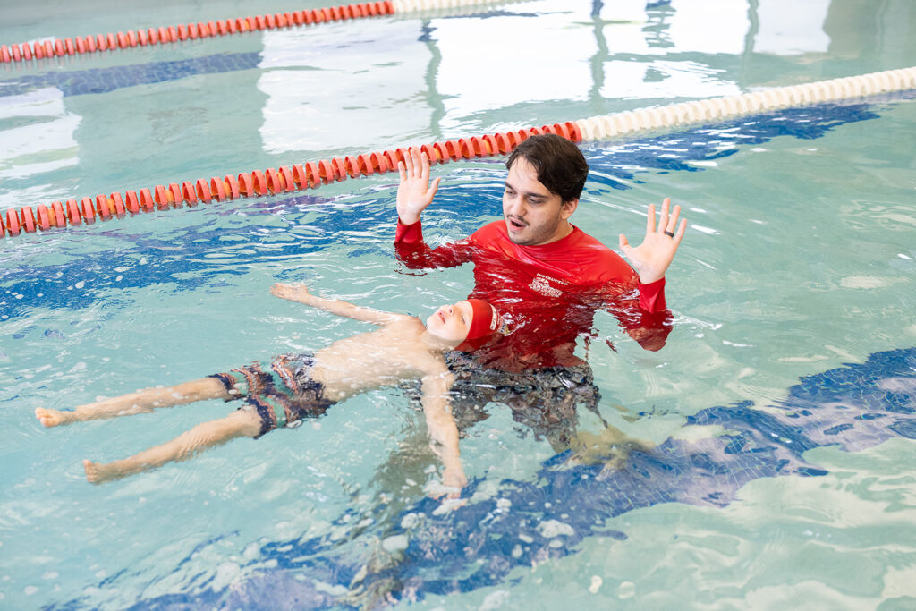 British Swim School instructor supporting a young child in a survival back float during an indoor swim lesson.