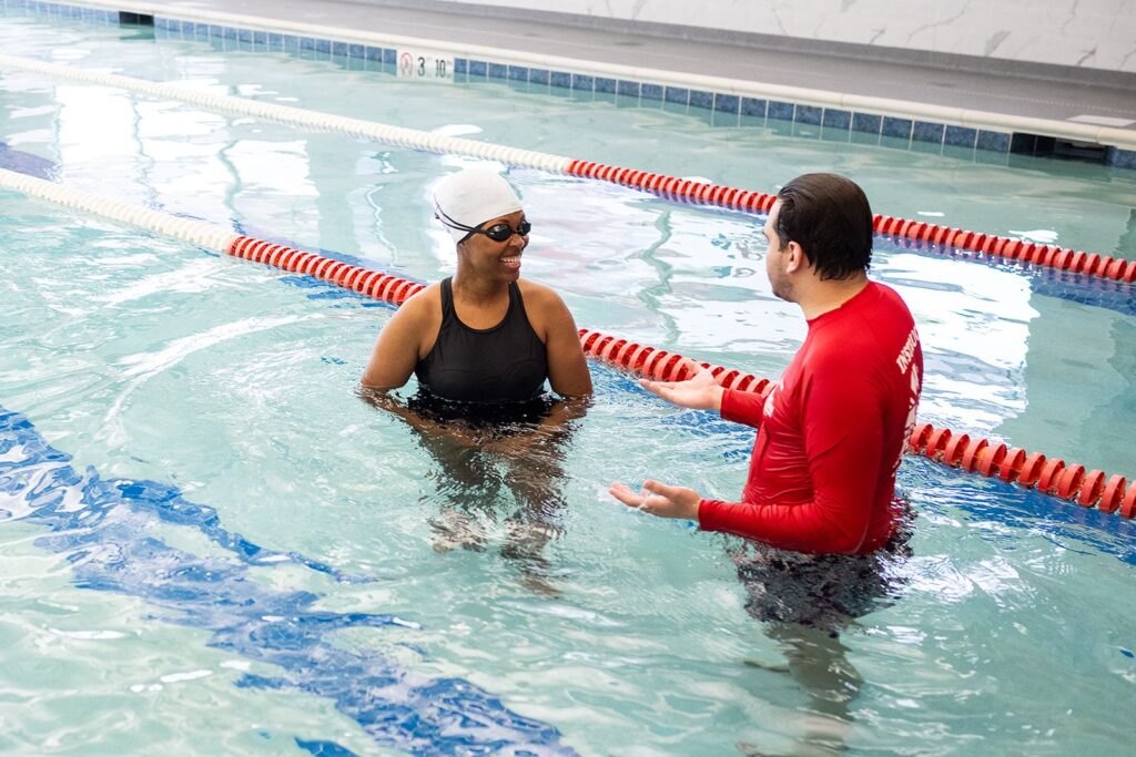 Adult woman in a white swim cap and goggles chatting with a British Swim School instructor in an indoor pool during a swim lesson in North Colorado Springs.