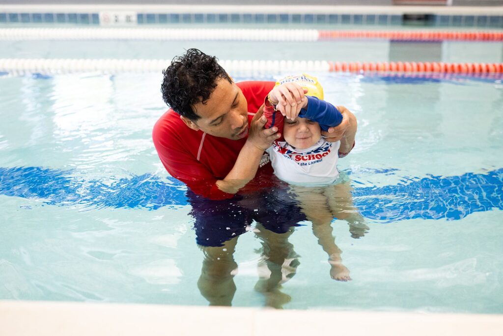 British Swim School instructor gently guiding a young child through water safety and stream-line position in an indoor pool.