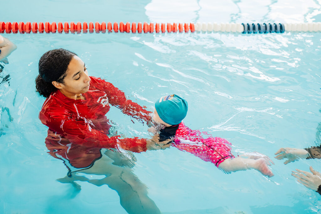 Swim instructor teaching a toddler foundational water competency and swimming skills in a pool.