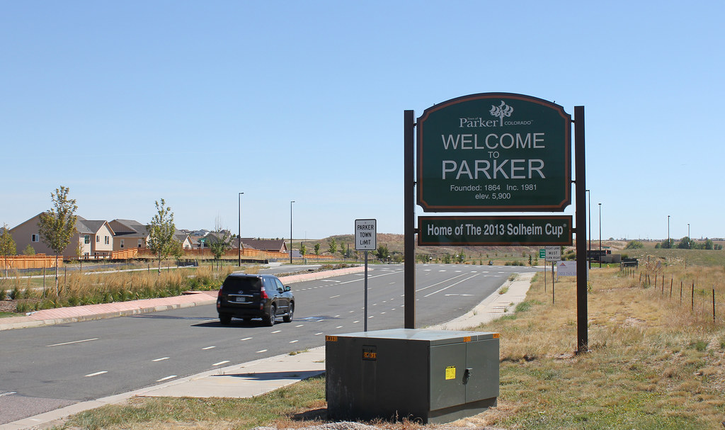 A Welcome to Parker sign in Colorado, displaying the town's founding in 1864 and its role as home of the 2013 Solheim Cup, situated next to a wide suburban road with new residential housing in the background under a clear sky.
