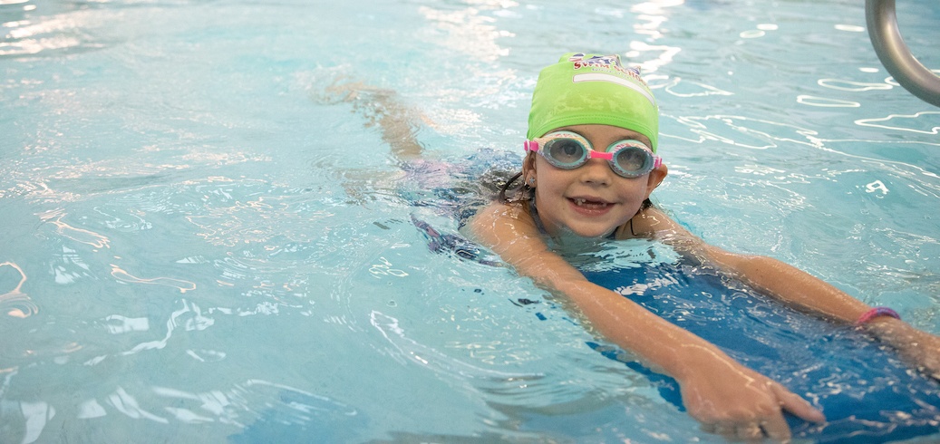 Smiling girl in a green swim cap and goggles practices swimming with a blue kickboard