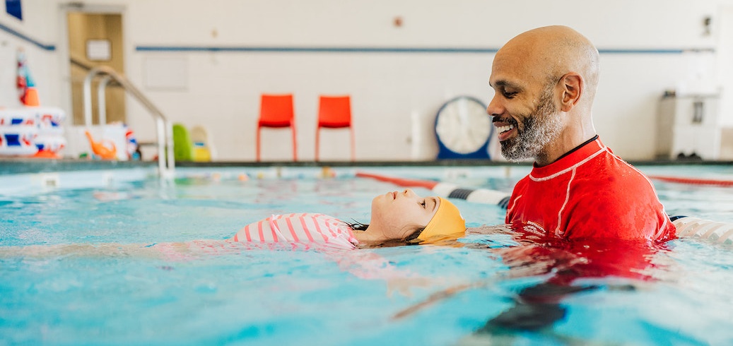Smiling swim instructor helps a young girl float on her back in a swimming pool.