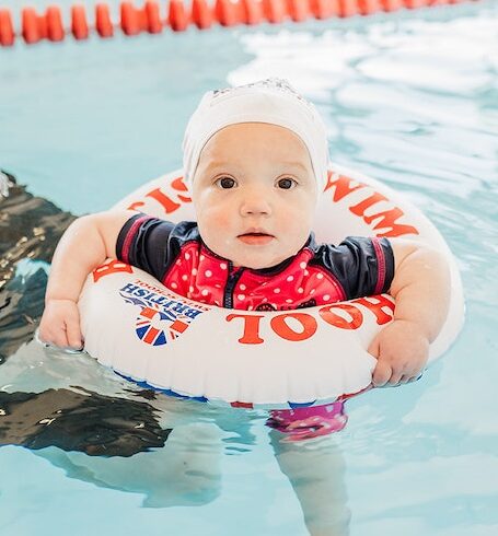 A baby in a white swim cap and red polka-dot rash guard floats in a British