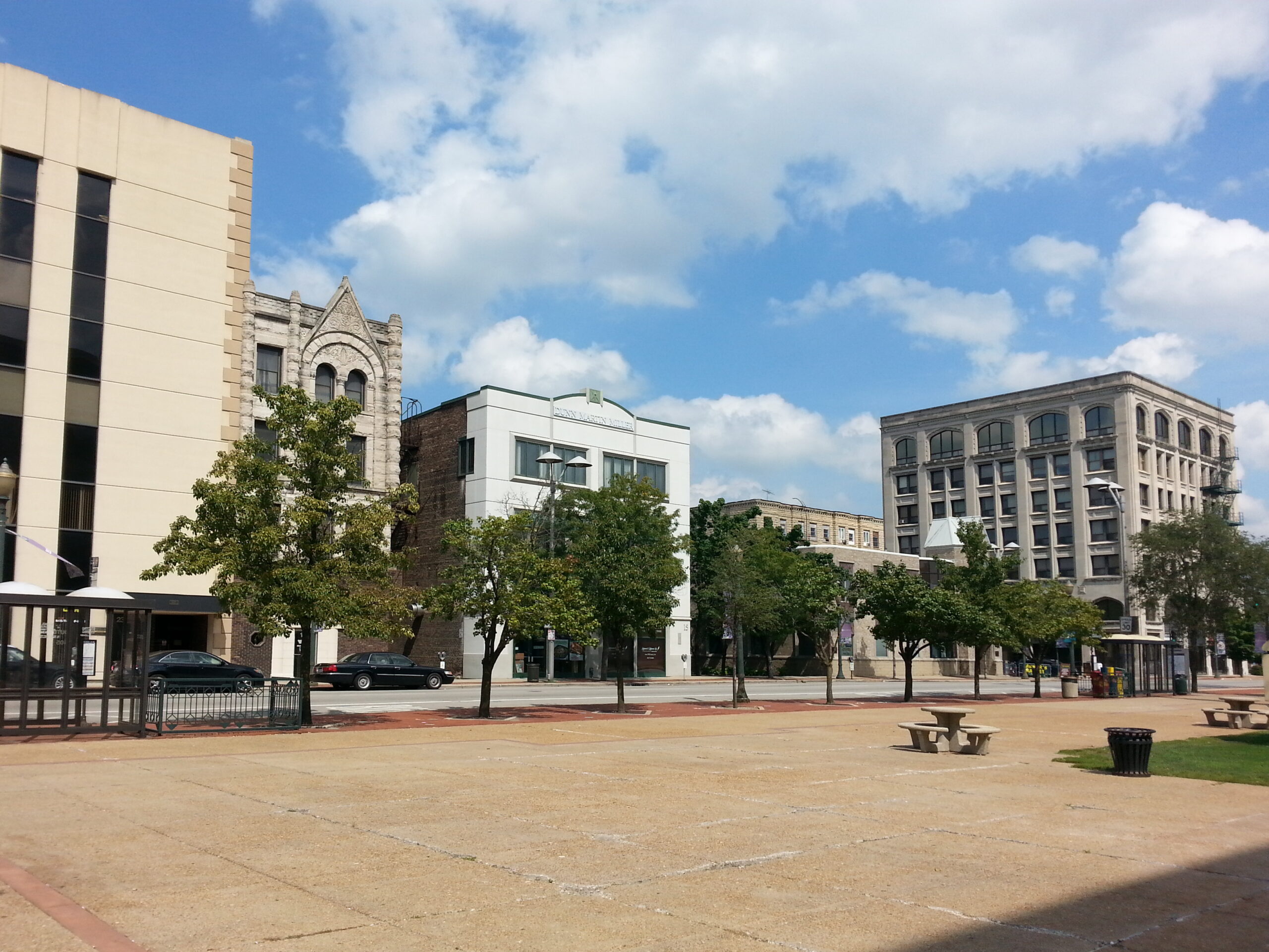 A sunny view of downtown Joliet, Illinois, featuring historic multi-story stone and brick buildings along a city street, a wide concrete plaza with park benches, and green trees under a bright blue sky with white clouds.