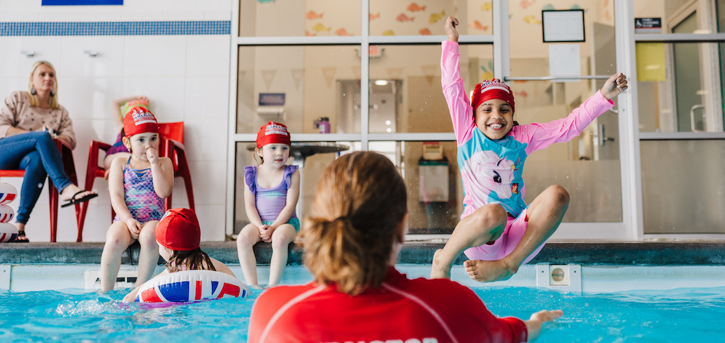 A young girl joyfully jumps into a swimming pool towards an instructor during a lesson.
