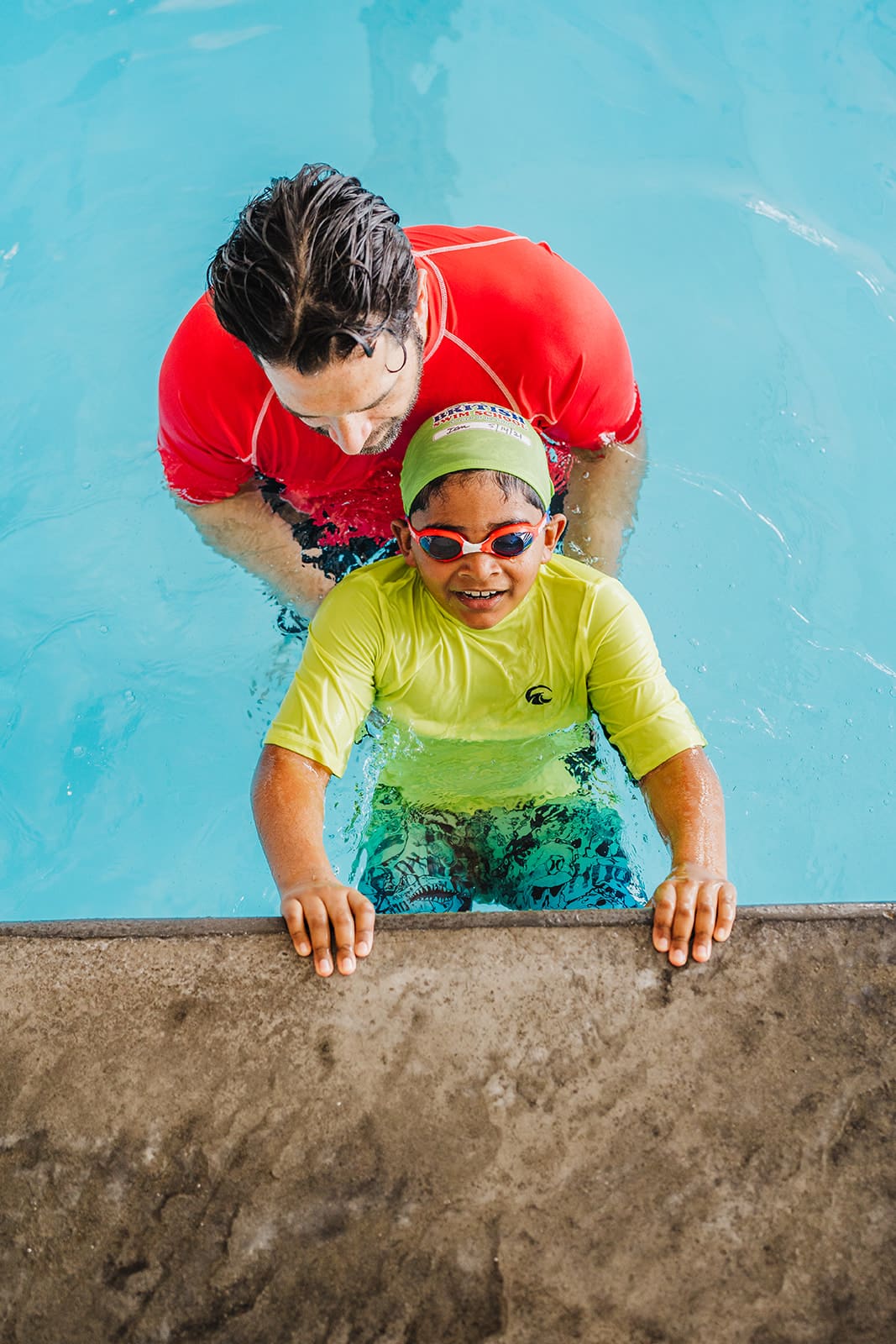 An adult instructor helps a smiling child in a green cap and goggles at the pool's