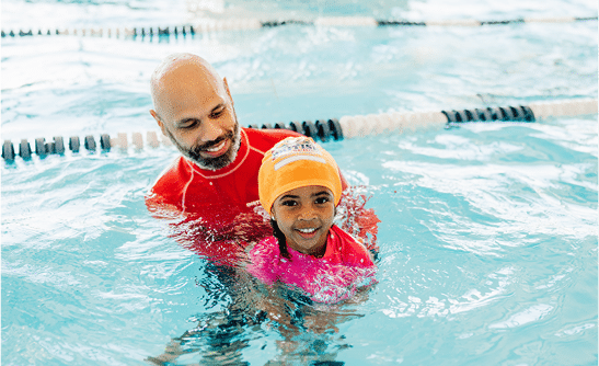 A smiling man in a red shirt teaches a young girl in an orange swim cap to