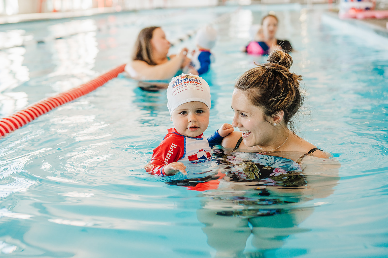 A smiling woman holds a baby in a Puddle Ducks swim cap during a swimming