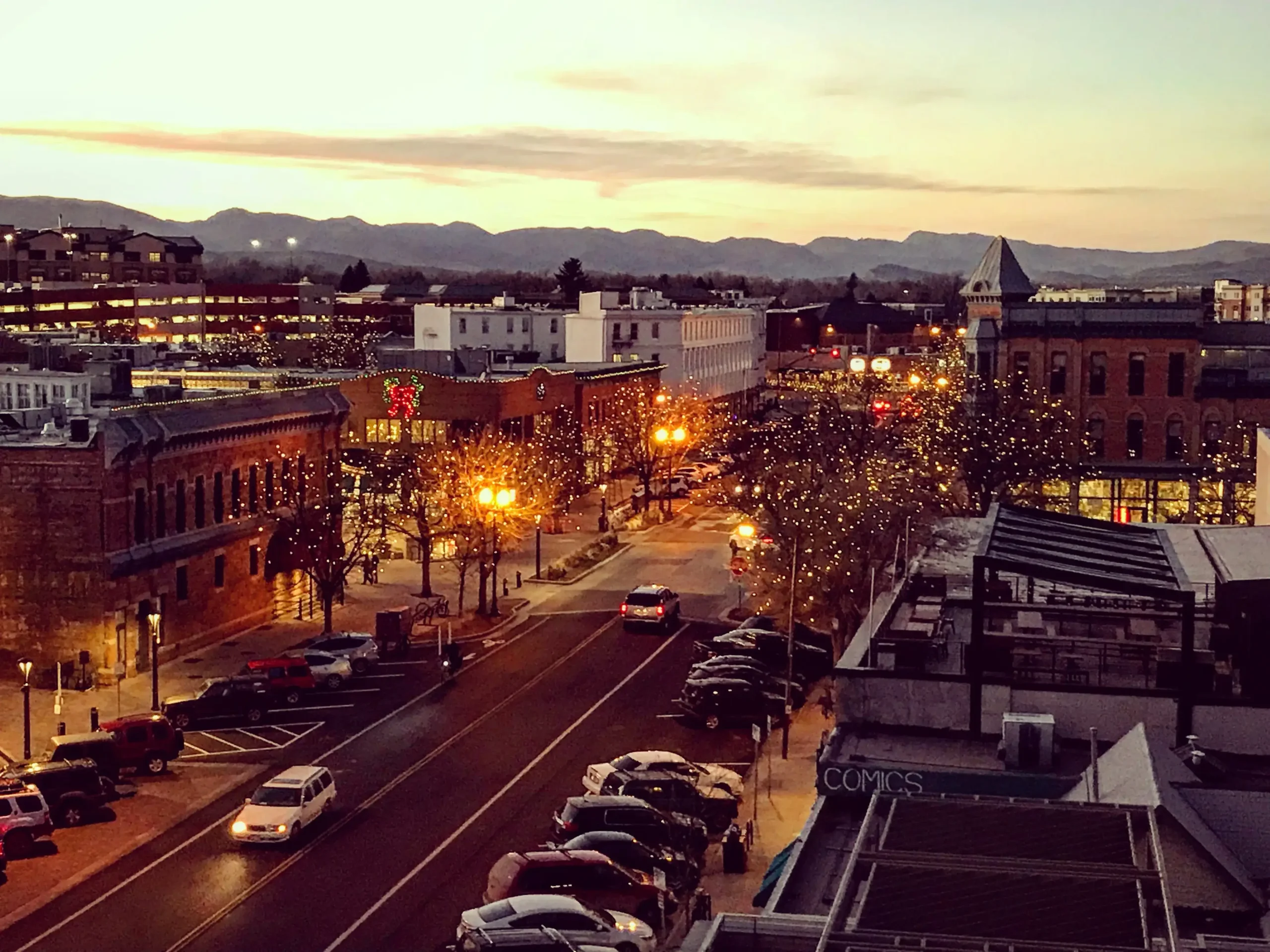 An aerial view of the historic downtown Old Town area of Fort Collins, CO at dusk, featuring Mountain Avenue lined with festive holiday string lights on trees and the scenic Rocky Mountain foothills in the distance.