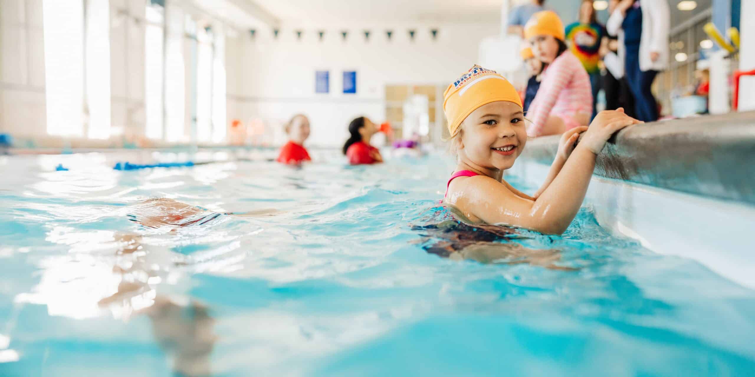 A smiling young girl in a yellow swim cap holds onto the edge of an indoor swimming pool