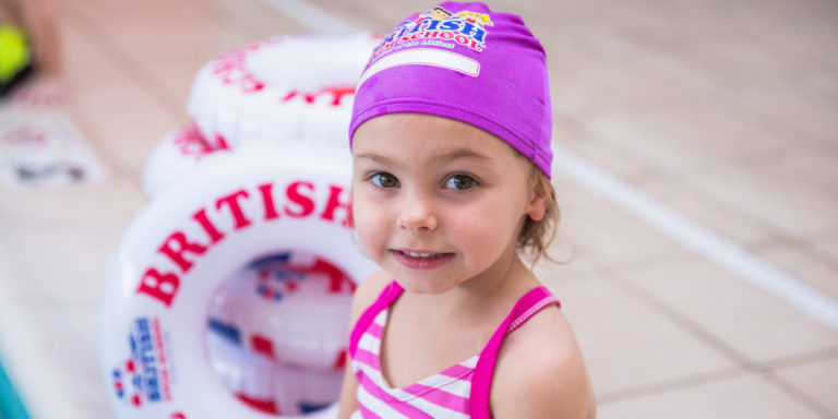 Little girl in a British Swim School swim cap smiling next to a pool