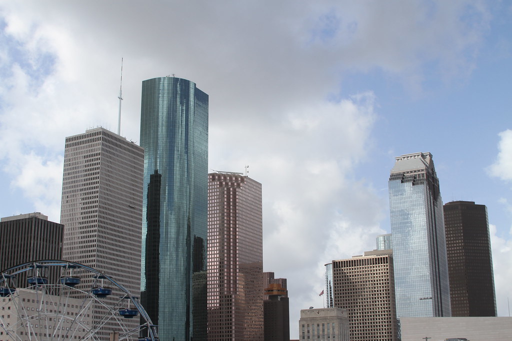 An urban skyline view of downtown Houston, Texas, featuring the green-glass Wells Fargo Plaza, the JPMorgan Chase Tower, and other high-rise skyscrapers with the top of a Ferris wheel visible in the foreground under a cloudy sky.
