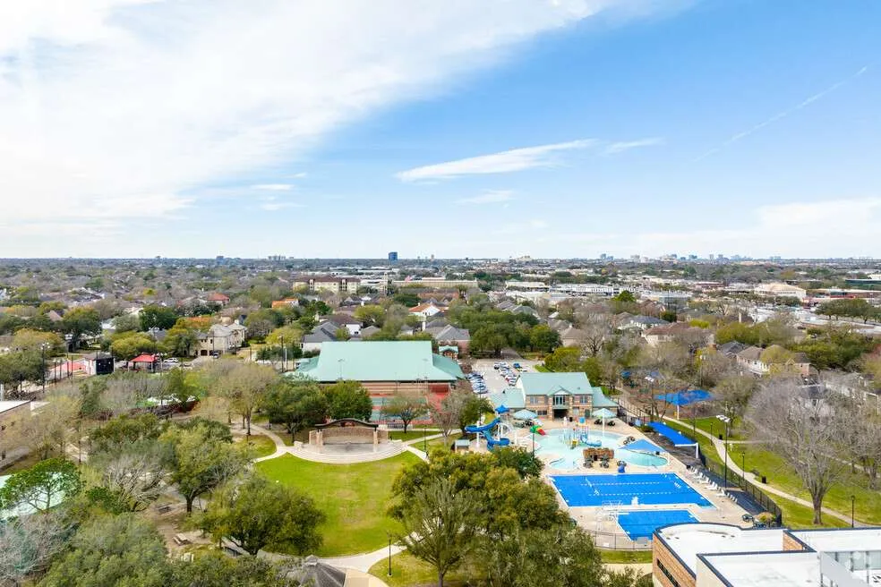 An aerial view of Bellaire Zindler Park in Bellaire, Texas, featuring the community pool with water slides, a large green lawn with an outdoor stage, tennis courts, and the surrounding suburban residential neighborhood under a bright sky.