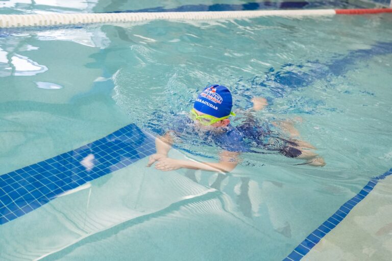 A child in a blue swim cap and yellow goggles swims in a clear pool.