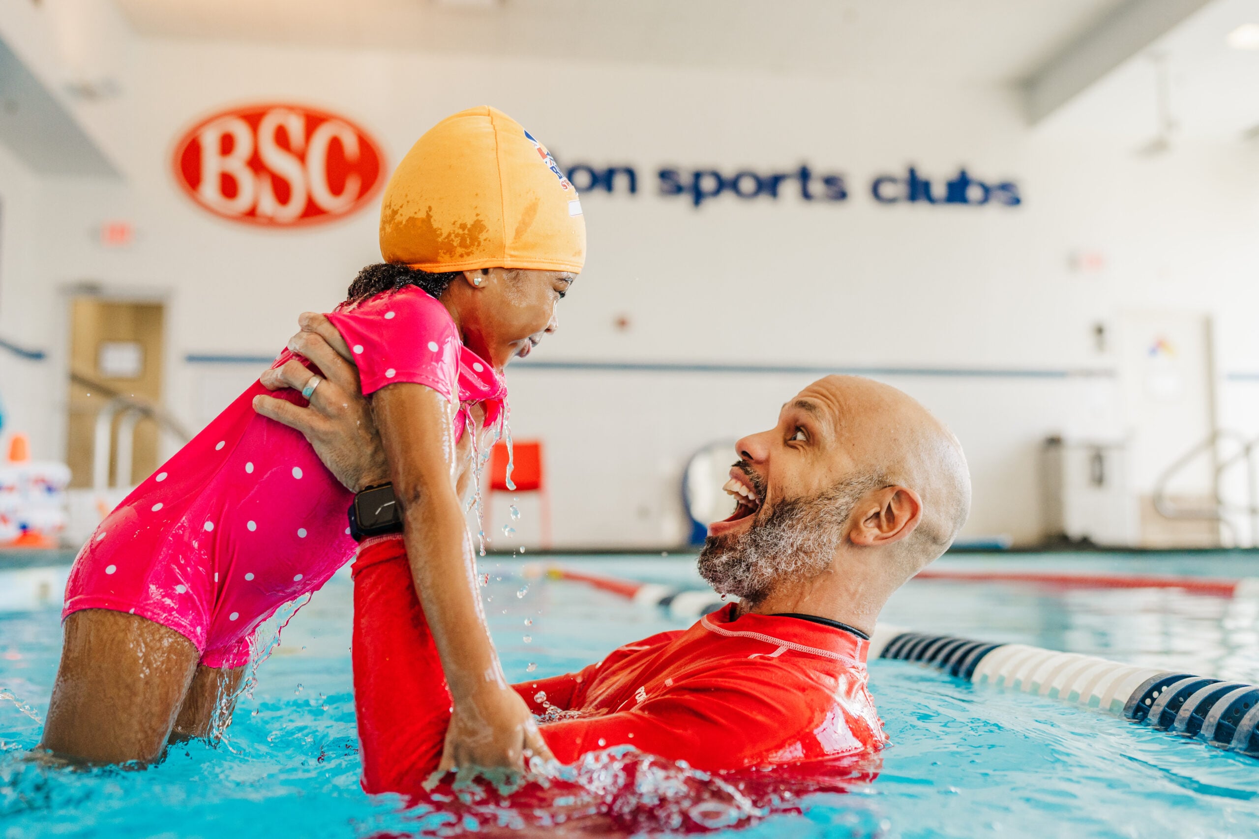 A happy instructor holds a smiling child in a bright indoor swimming pool.